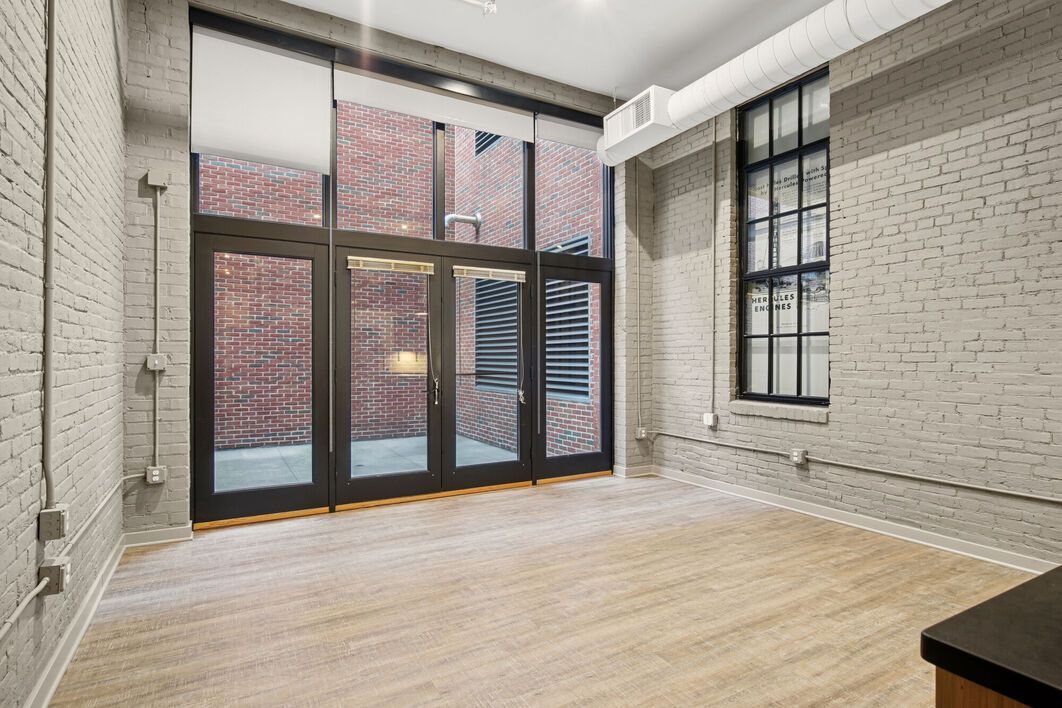 Empty room with large glass doors and brick wall view, wood floors, exposed brick walls, and black-framed windows.