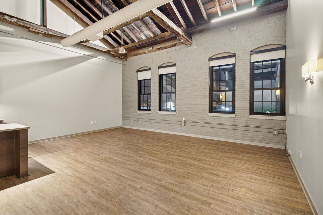 Empty, open-plan loft with exposed brick wall, three windows, hardwood floors, and high ceilings with exposed beams.