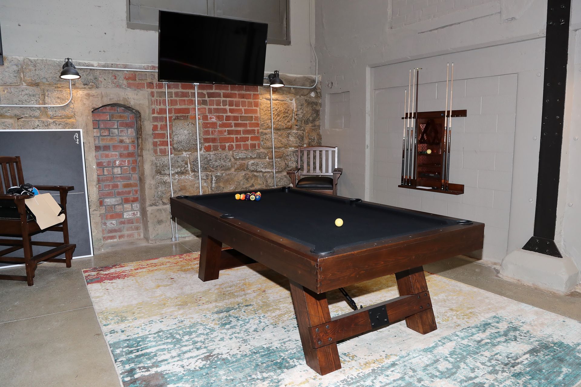 Pool table in a dimly lit room with brick wall and TV, rug, cue rack.