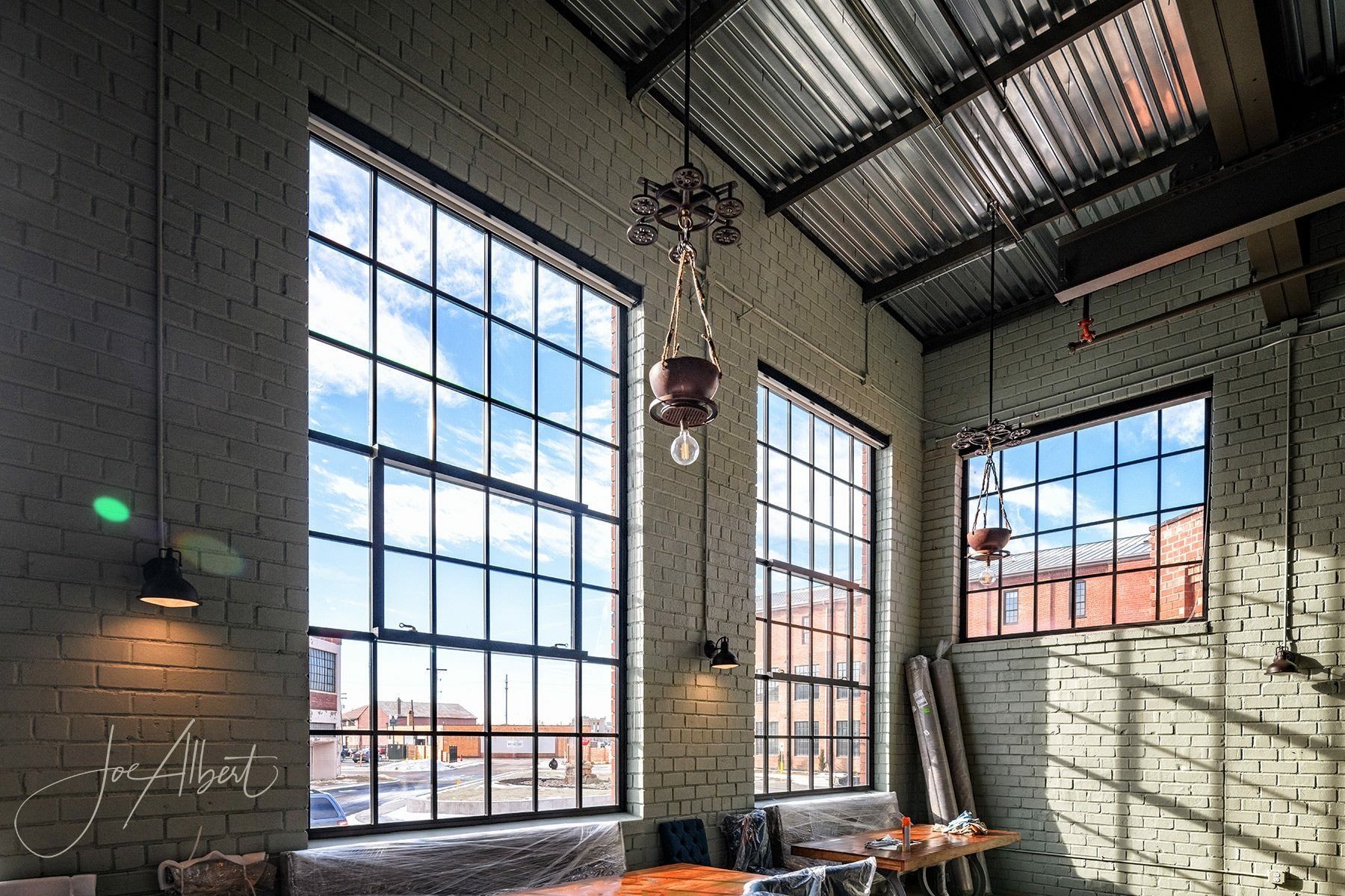 High-ceiling industrial interior with large windows, exposed brick, and metal roof. Sunlight streams in.