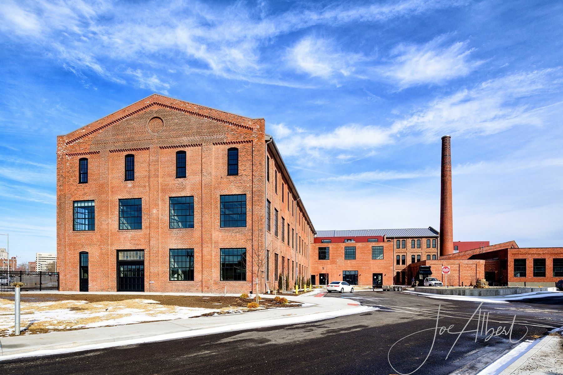 Brick building with large windows and tall smokestack against a cloudy blue sky.