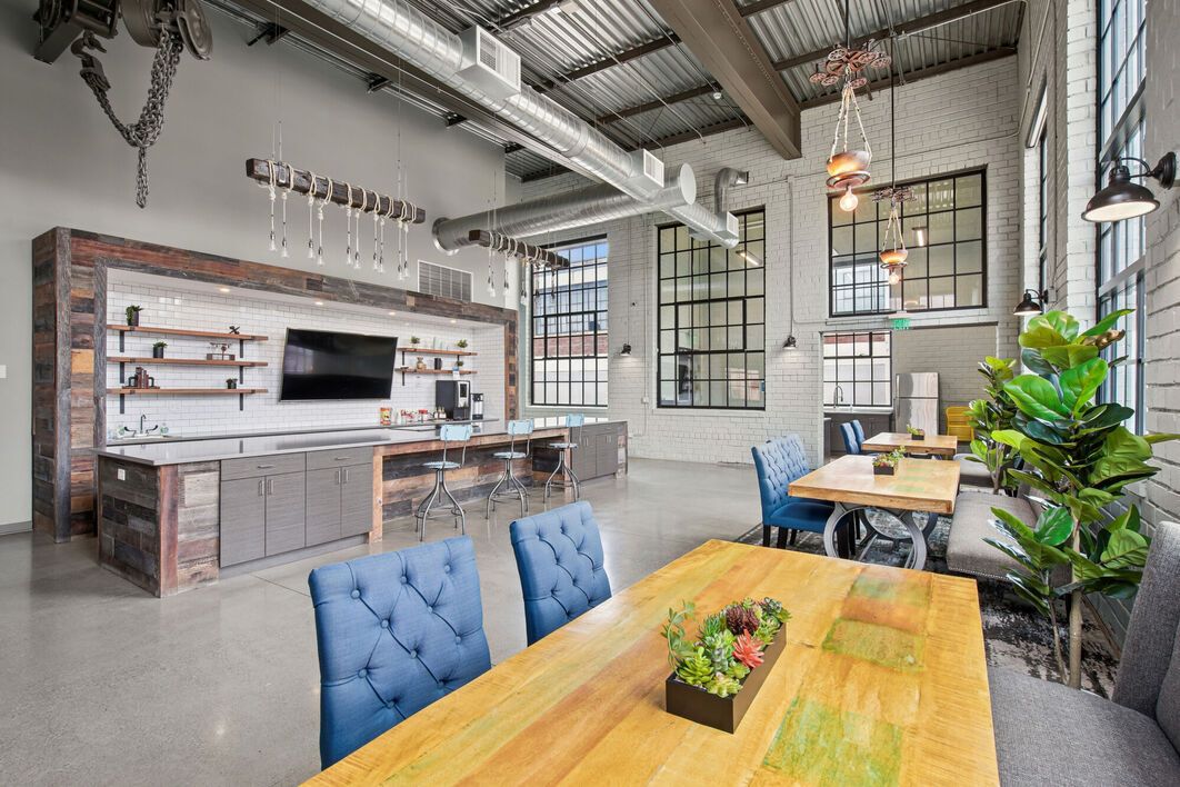 Modern industrial-style interior with a dining area featuring a long wooden table, blue chairs, and a built-in kitchen.
