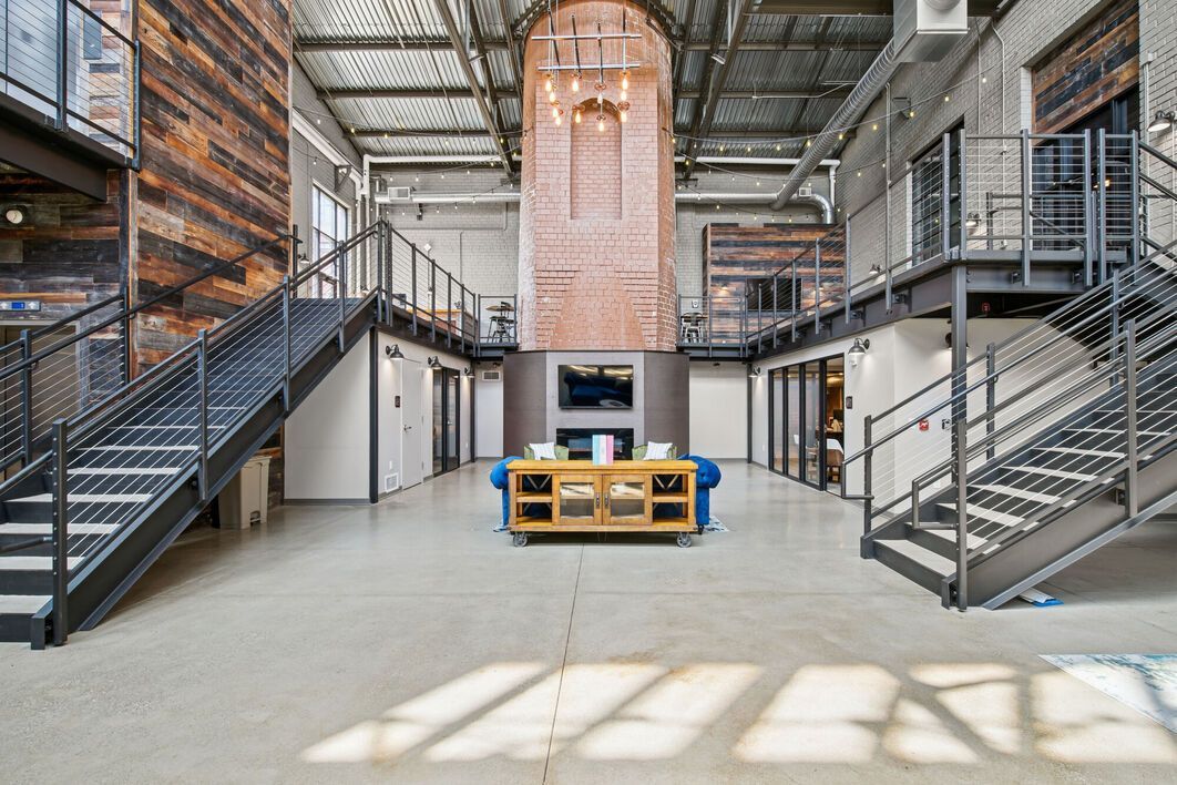 Large industrial-style interior with exposed brick, metal staircases, and concrete floor. Central sofa faces a fireplace.