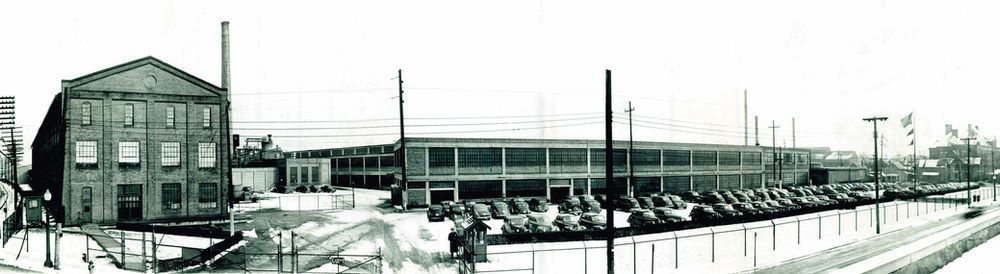 Black and white panoramic of industrial buildings and parked cars, possibly winter.