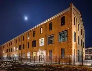 Brick building at night with lit windows, fence in front, moon visible in the dark sky.