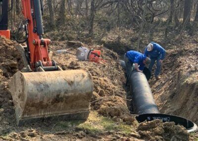 Workers installing a large pipe in a ditch with an excavator and chainsaw nearby.