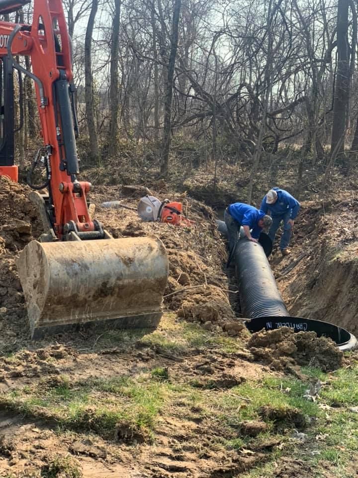 Two men installing black drainage pipe in a trench; orange excavator nearby in a wooded area.