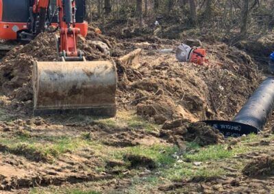 An excavator bucket digging dirt near a dark pipe on a muddy construction site.