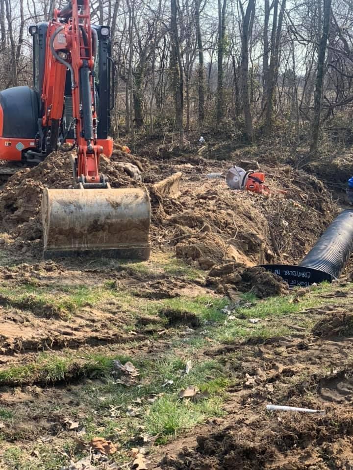 Orange excavator digging dirt next to a black pipe, in a wooded area.