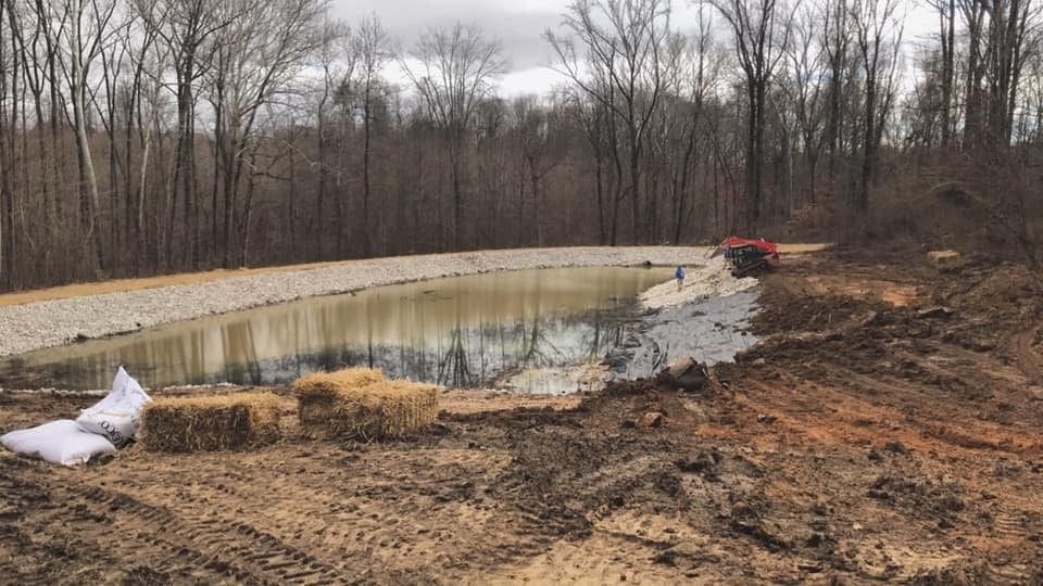 Pond with rocky edges, hay bales, and a small tractor in a wooded area.