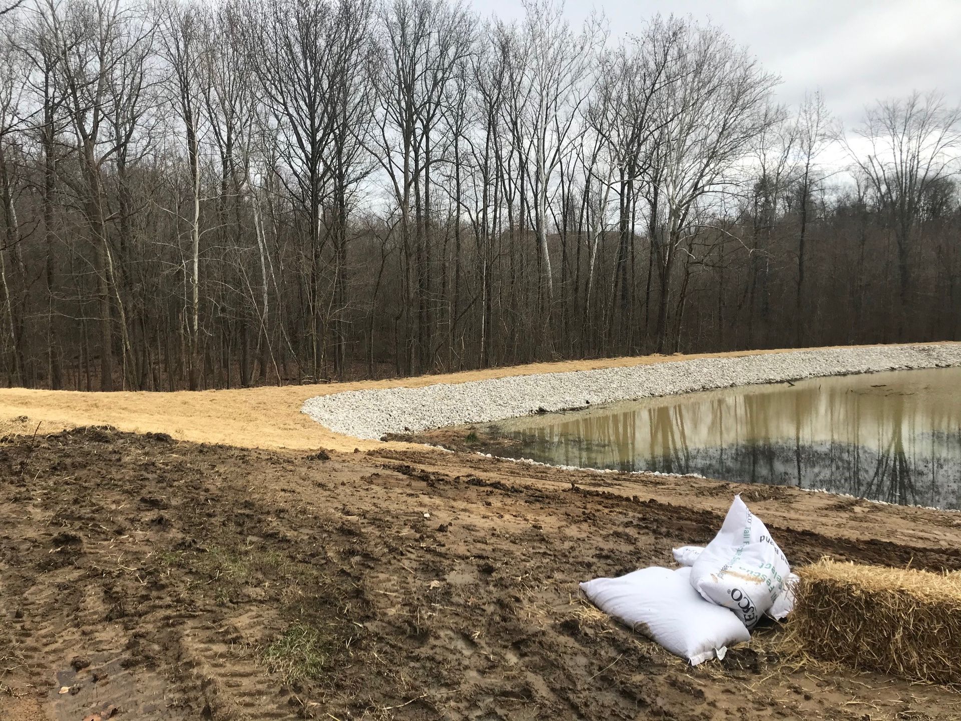 A muddy shoreline with a pond, trees, and two white bags.