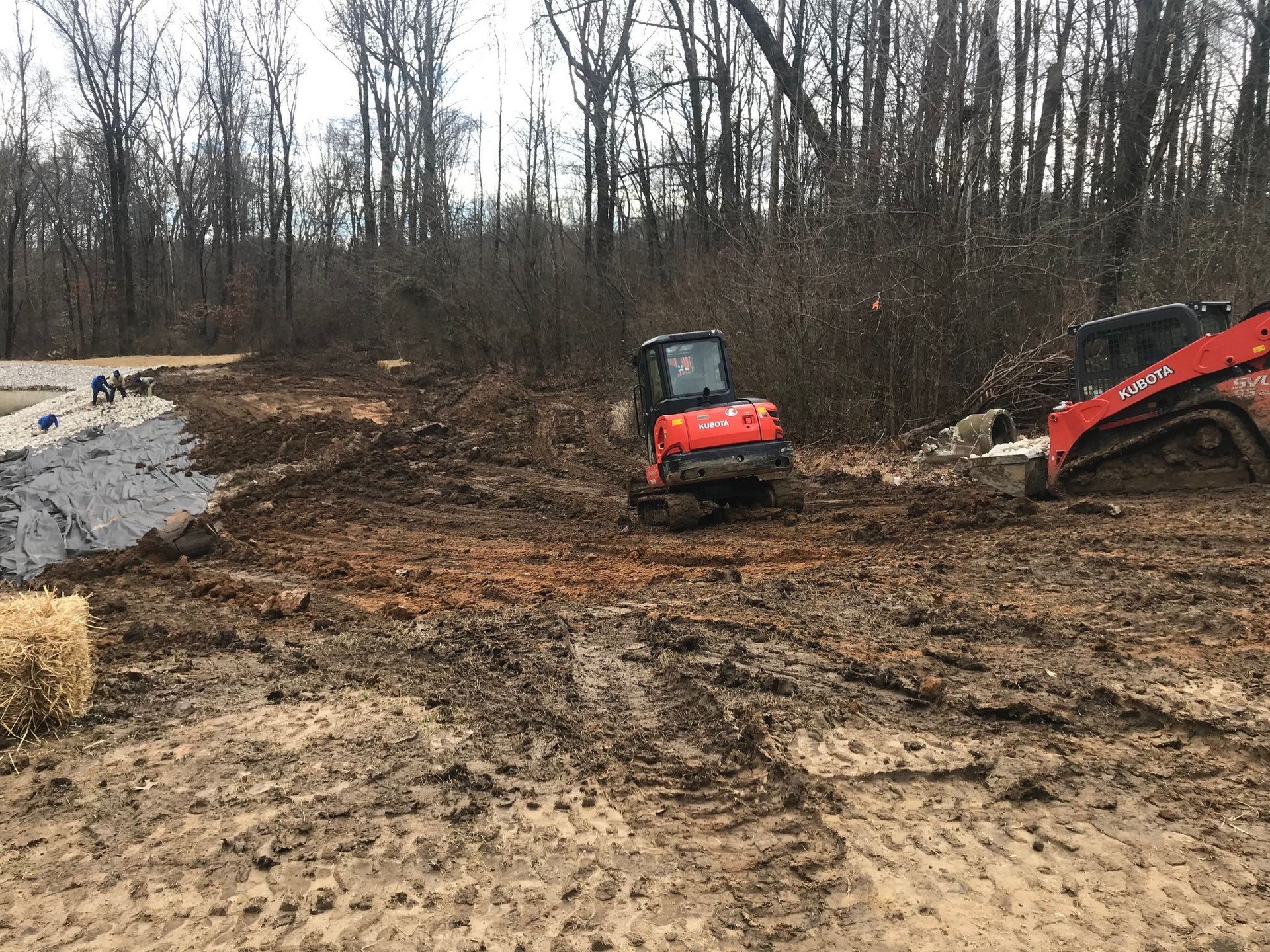 Muddy construction site with orange Kubota excavator and skid steer. Bare trees in background.
