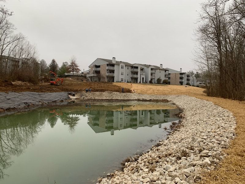 Pond with rocky shoreline reflects an apartment building under a cloudy sky; brown grass and bare trees surround it.