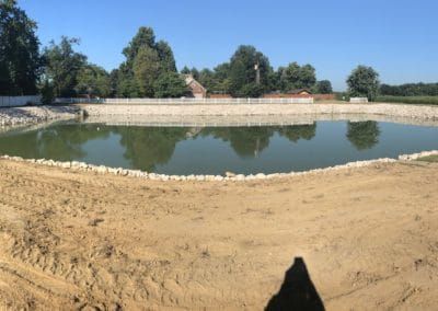 Pond with stone border surrounded by sandy earth, trees, and a building under a blue sky.
