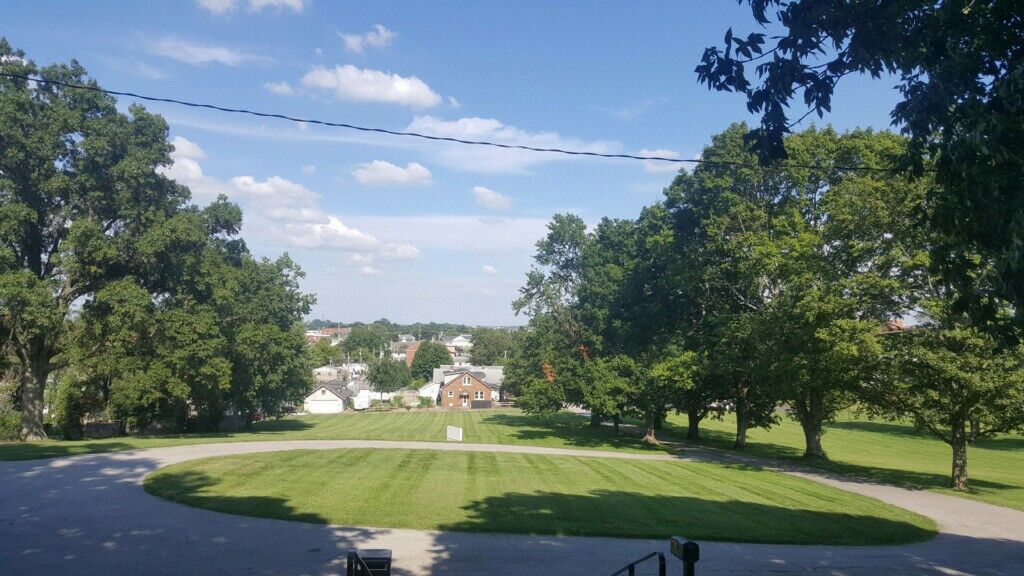 Scenic view of a grassy park with trees, a pathway, and a distant town on a sunny day.