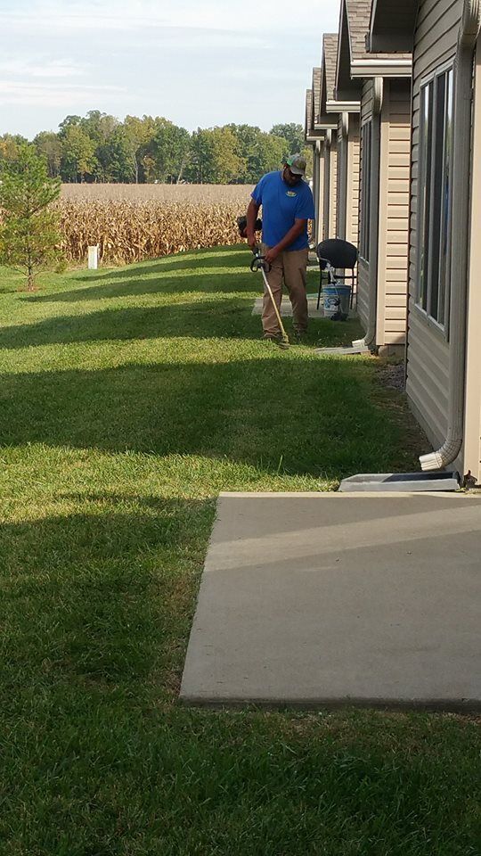 Man in blue shirt using a weed eater on a grassy lawn next to a house, with a field in the background.