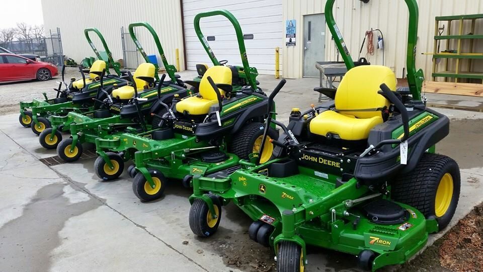 Five John Deere zero-turn mowers in a row, green and yellow, parked outside a building.