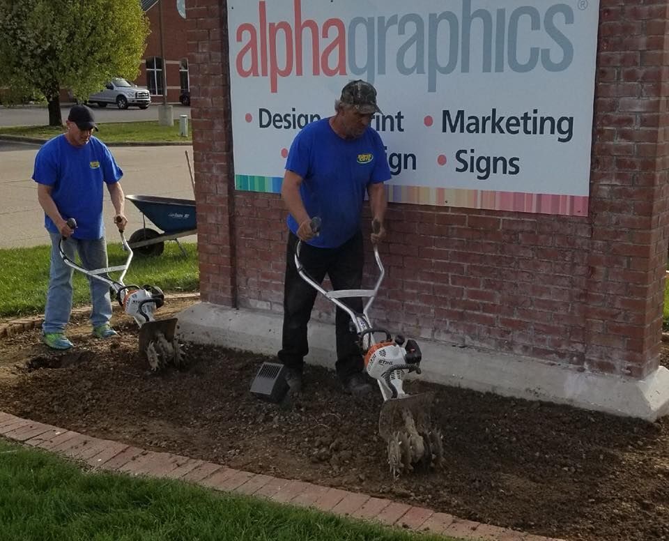 Two men tilling soil in front of an AlphaGraphics sign. They are wearing blue shirts and using tillers.