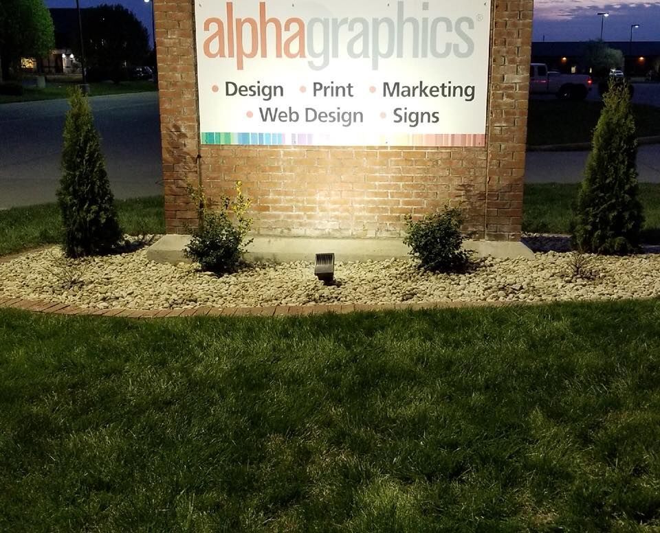 Sign for AlphaGraphics with services listed, fronted by grass and landscaping.