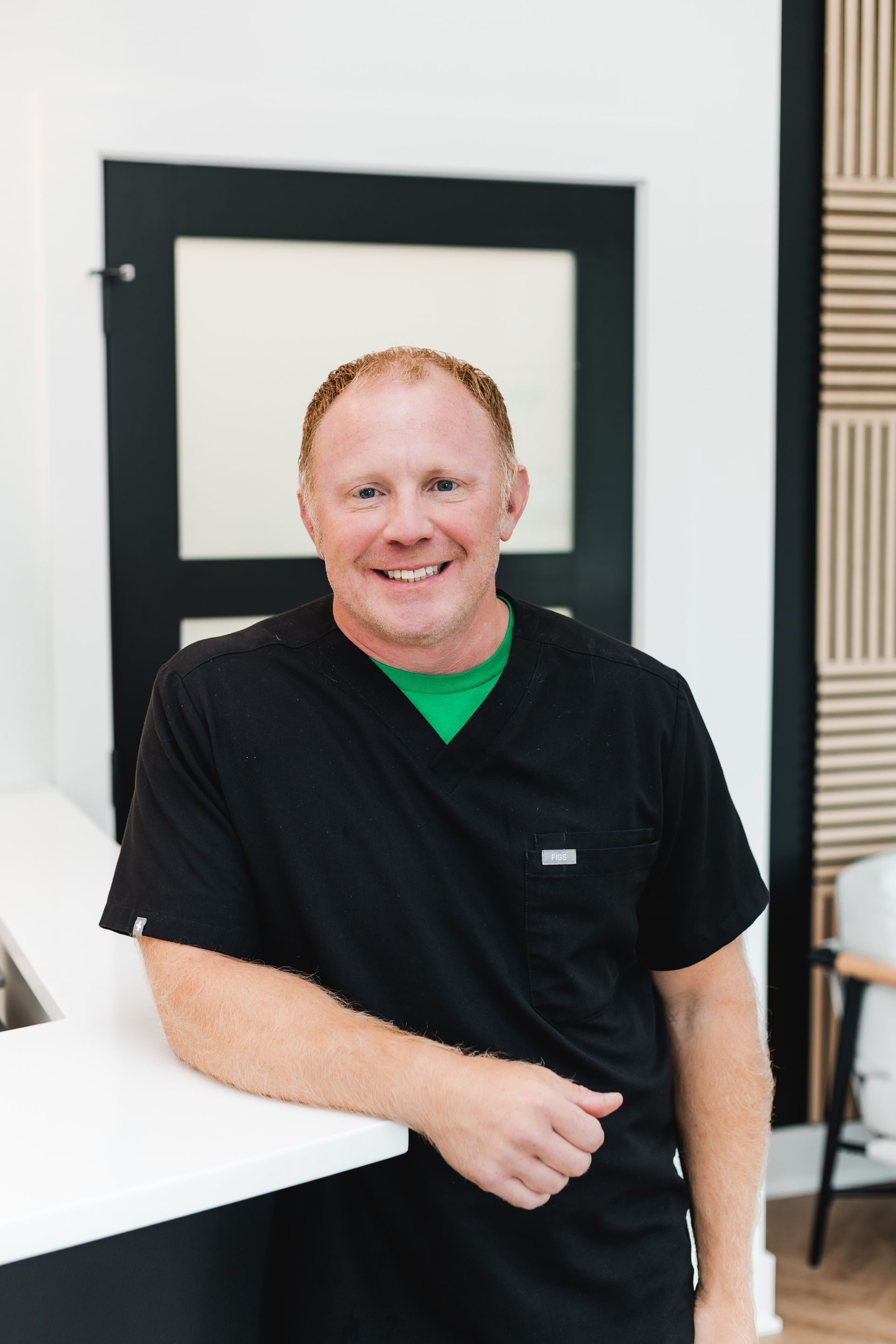 Man in black scrubs, green undershirt smiles, leaning on white counter, near black door, and beige wall accent.