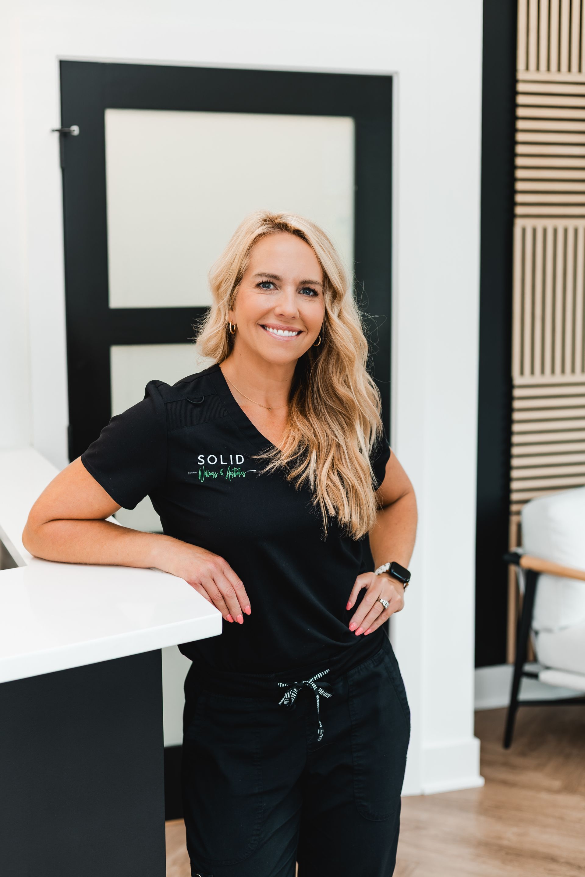 Woman in black scrubs, smiling, leaning on a white counter. Interior setting, door and chair in background.