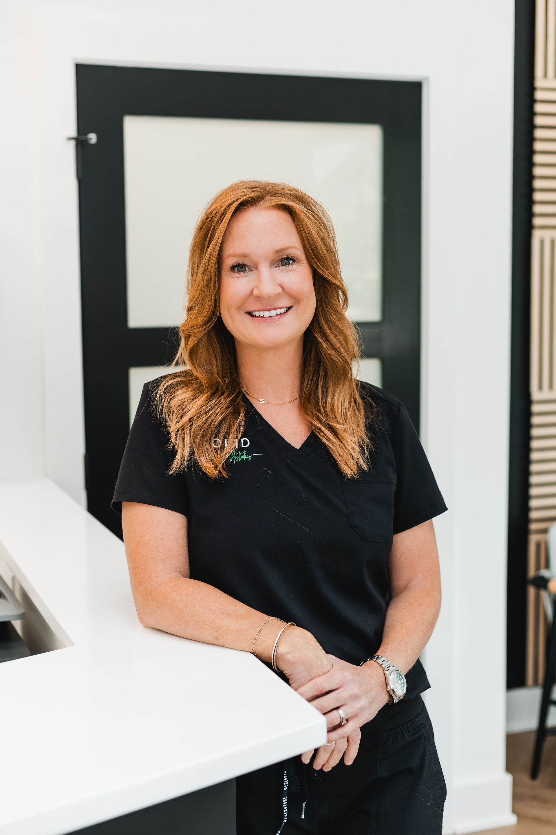 Woman with red hair in black scrubs smiles, leaning on white counter. Door in background.