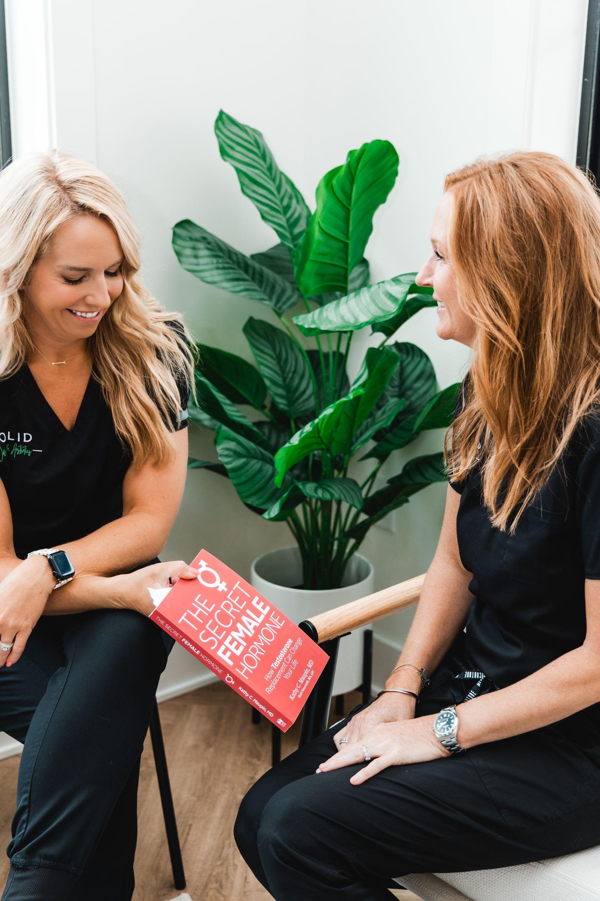 Two women in black scrubs seated, looking at a red book titled