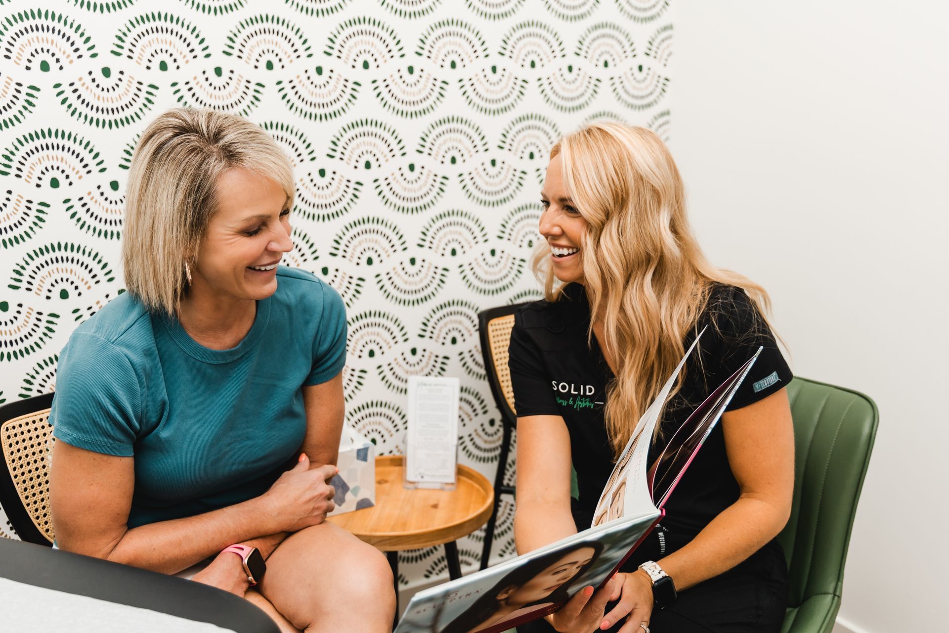 Two women laughing, one holding a brochure, in a room with patterned wallpaper.