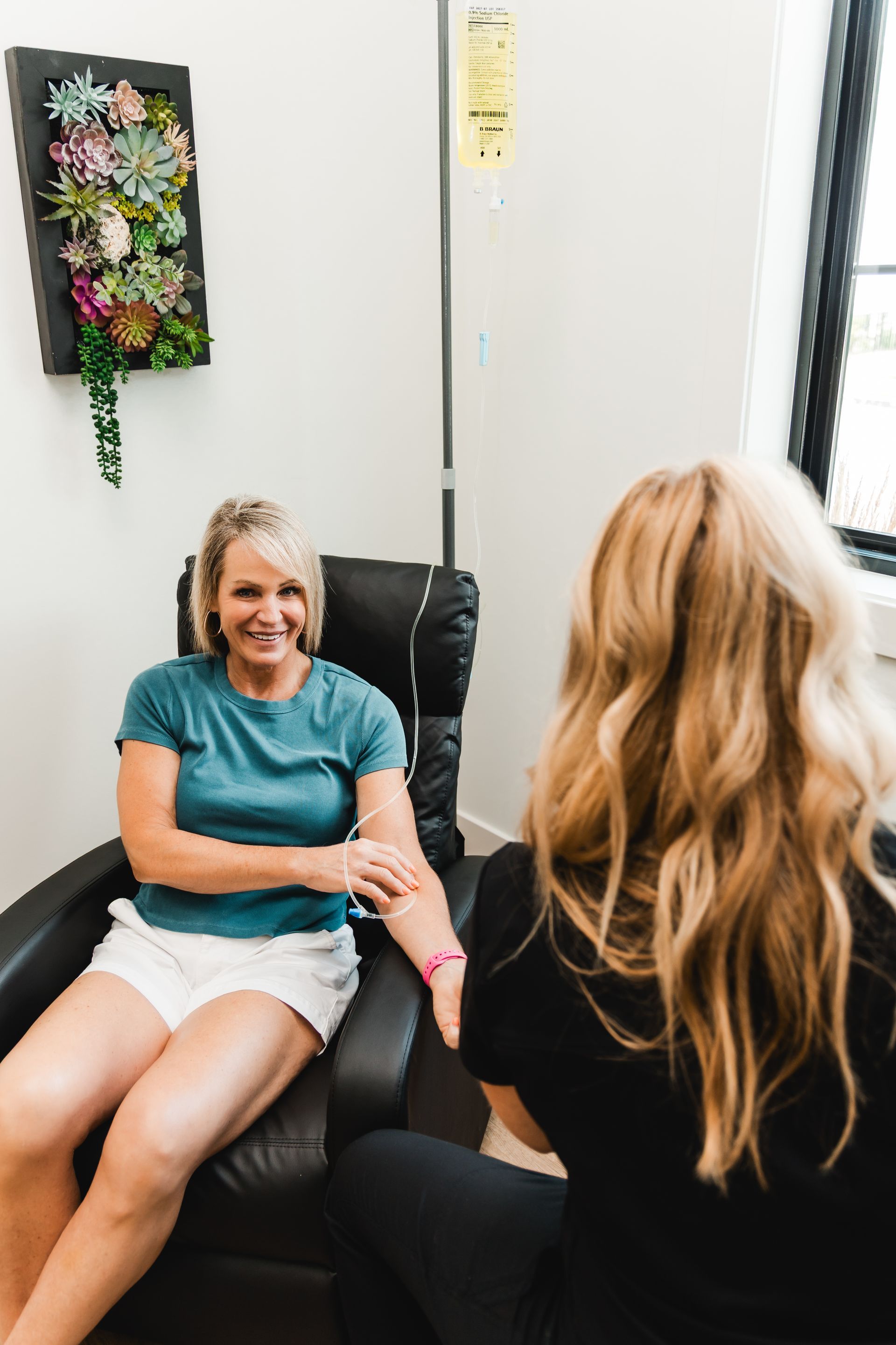 Woman receiving IV therapy, sitting in a black chair. Nurse monitors her, hand on patient's. Bright room, plant art.