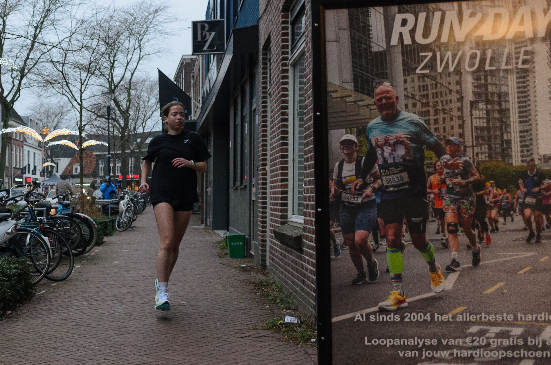 Een vrouw rent over de stoep naast een poster die reclame maakt voor Run Day Zwolle, in een stadsstraat.