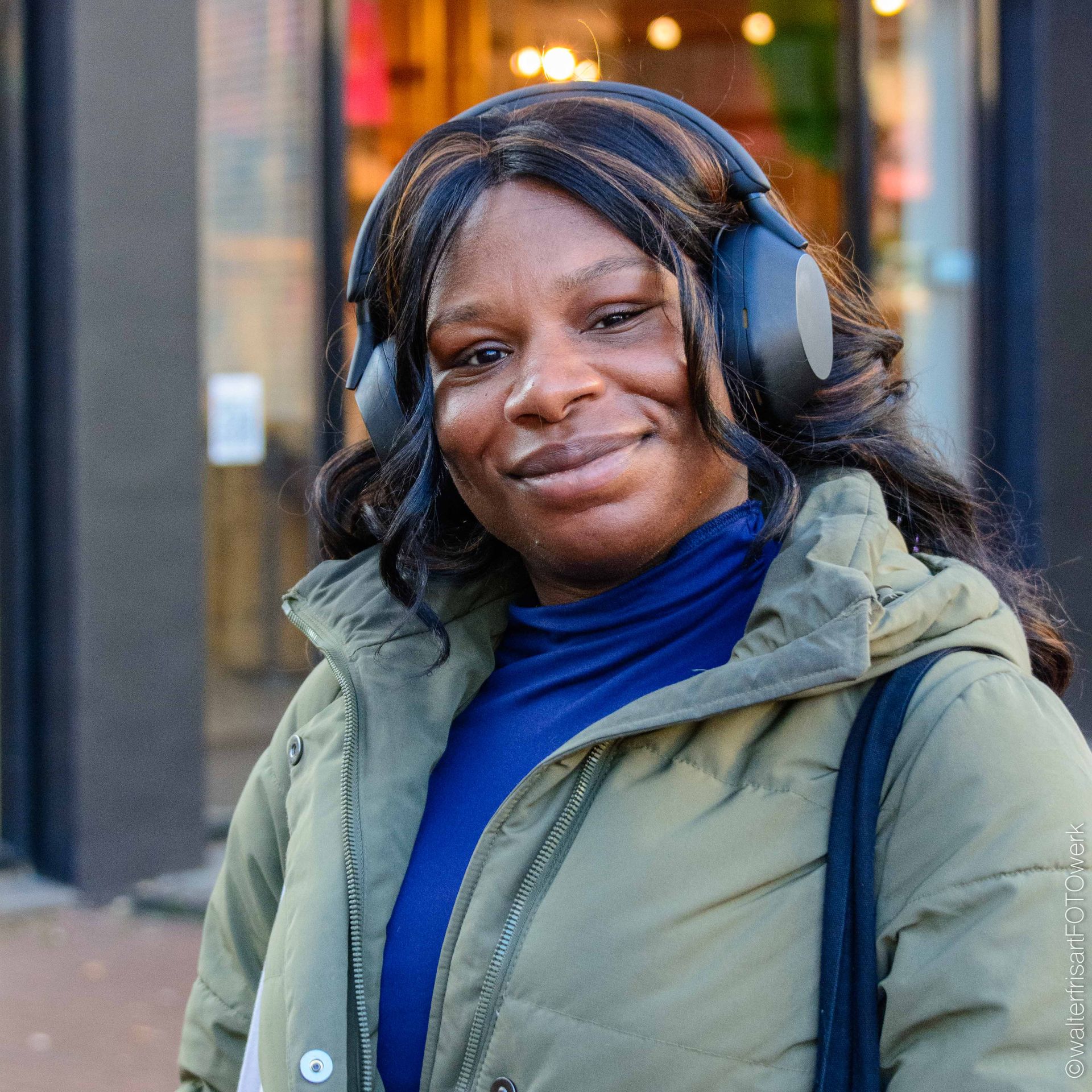 Woman wearing headphones and a green coat, smiling outside a building.