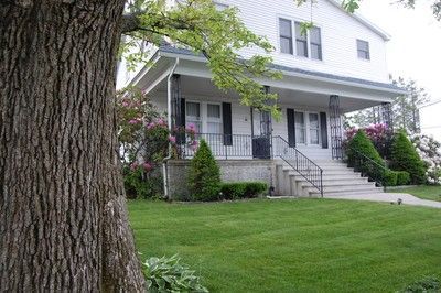 White two-story house with front porch, steps, and green lawn. A large tree trunk is in the foreground.