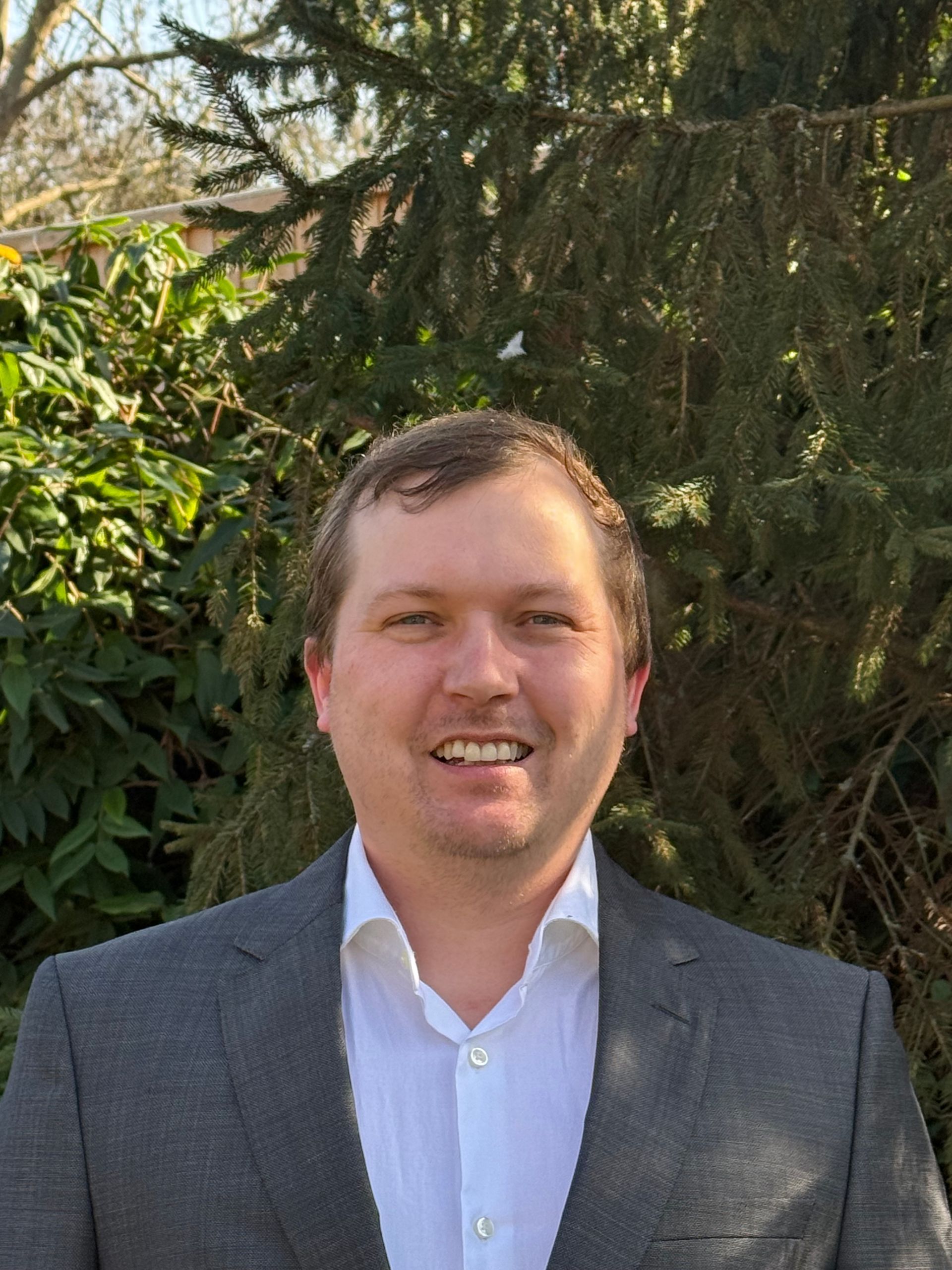 Photo of Jaime in a charcoal suit jacket and white shirt, standing in front of a tree.