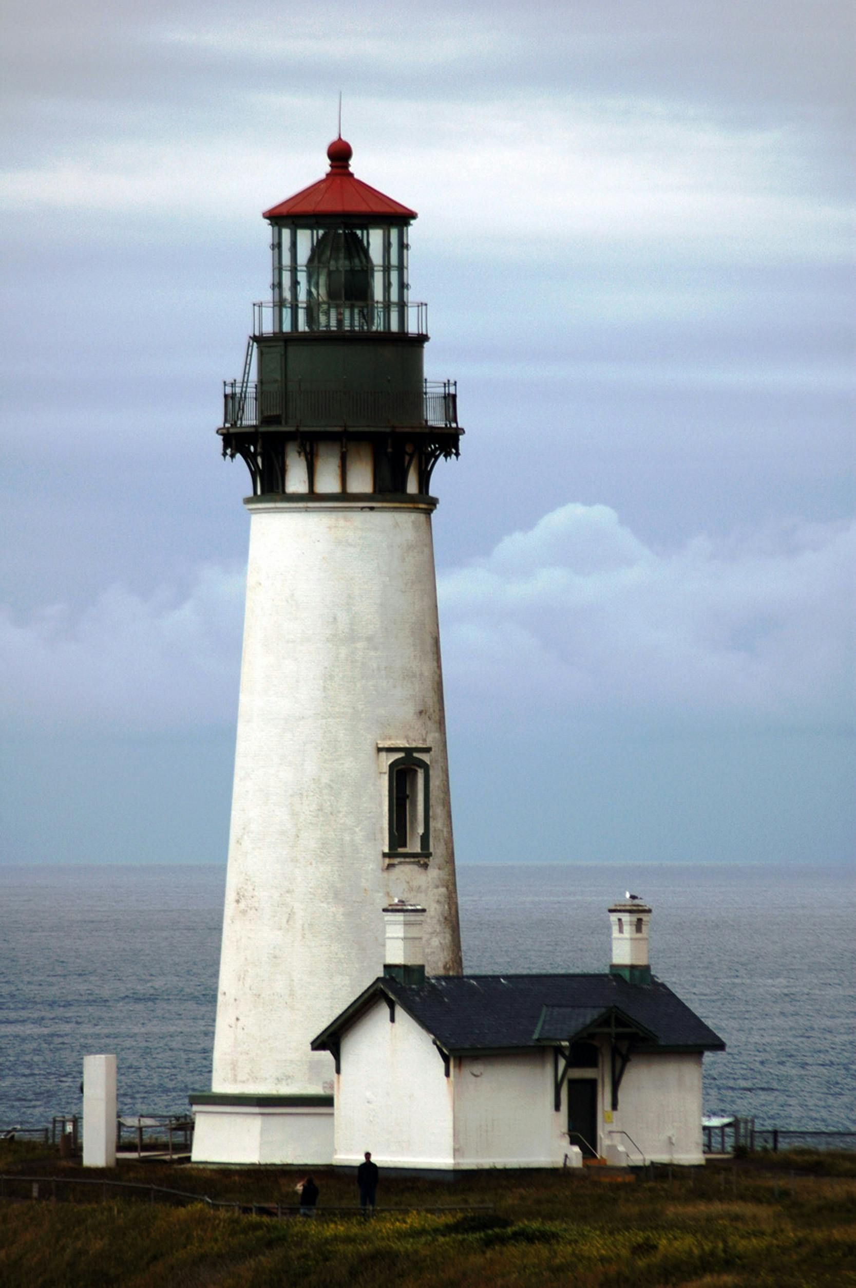 White lighthouse with red top and attached building, overlooking the ocean under a cloudy sky.