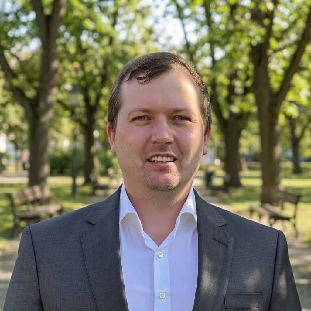 Photo of Jaime in a charcoal suit jacket and white shirt, standing in front of a tree.