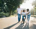Adult and two children ride bicycles along a sunny park path, one child learning with help.