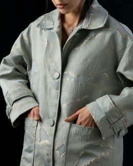 Woman in a long, light blue coat with gold embellishments, hands in pockets, against a black background.