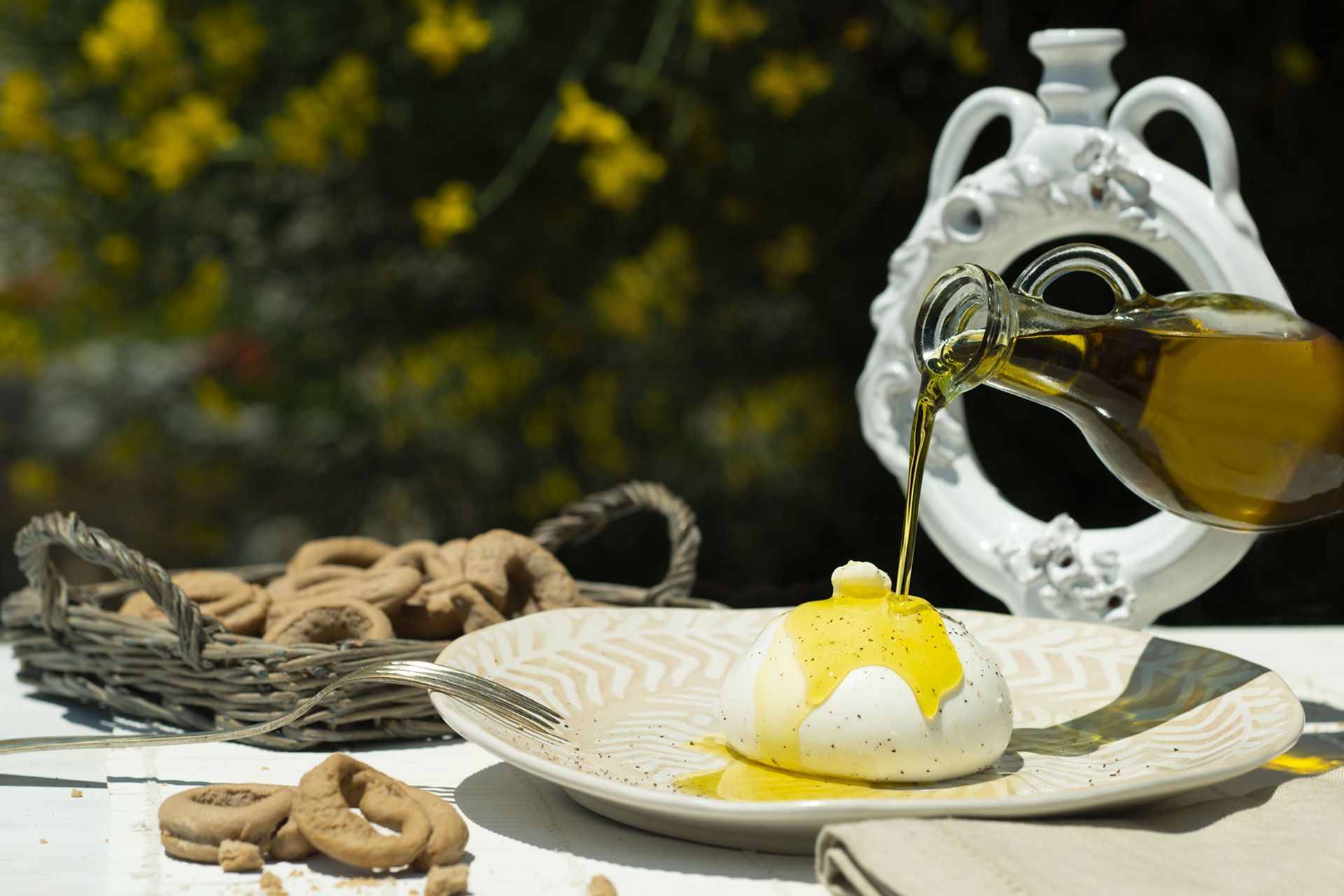 Olio d'oliva versato su una pallina di burrata su un piatto, cestino di biscotti, vaso bianco, all'aperto.