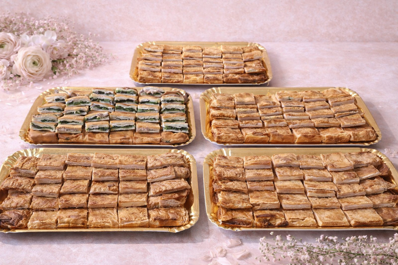 Cinque vassoi di pasticcini rettangolari su uno sfondo rosa chiaro, decorati con fiori.