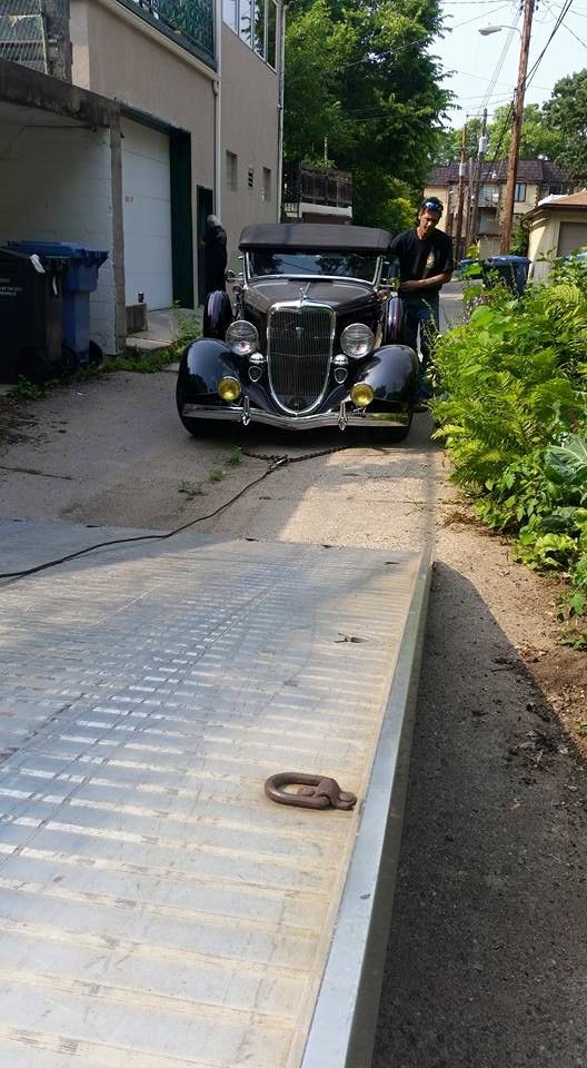 A black vintage car on a flatbed tow truck in an alley, with a man standing nearby.