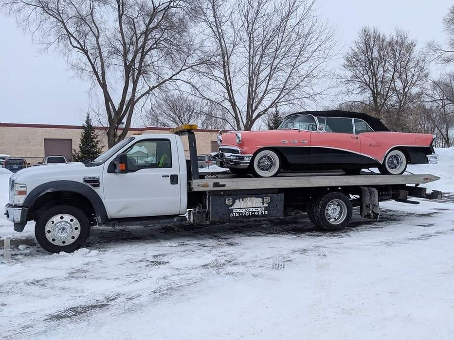 White tow truck carrying a pink and black classic car in a snowy setting.