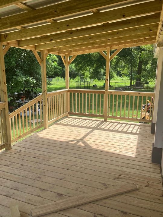 A newly constructed wooden deck with a covered roof, railings, and staircase overlooking a grassy yard with trees.