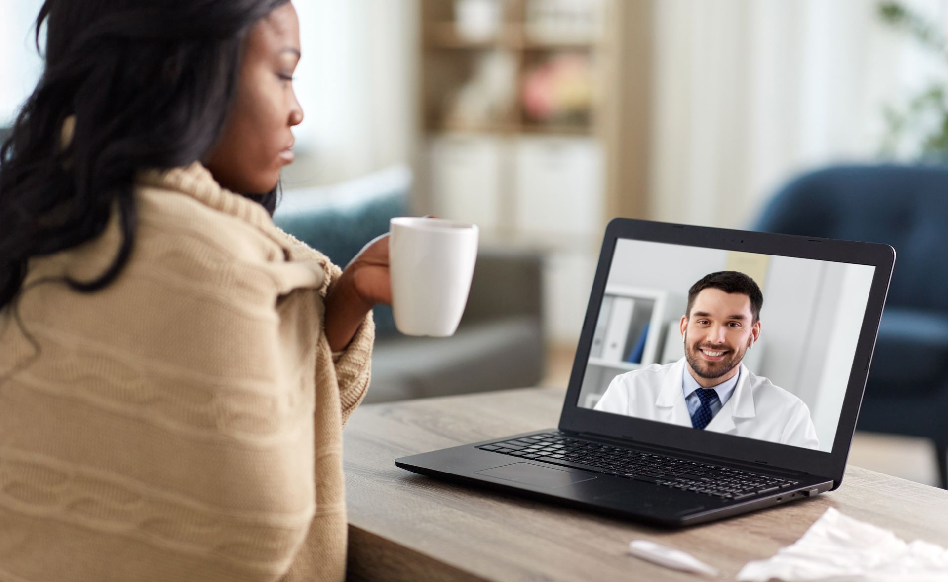 A woman is sitting at a table with a cup of coffee and talking to a doctor on a laptop.