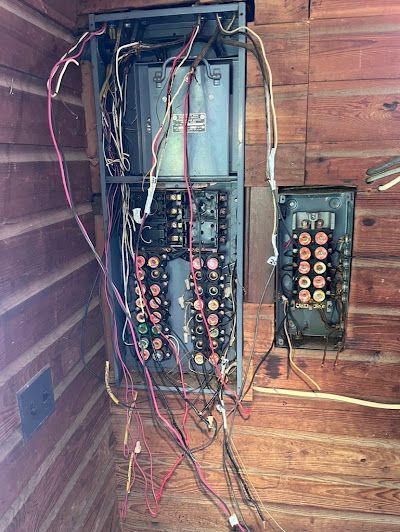 A very messy electrical panel is sitting on a wooden wall.
