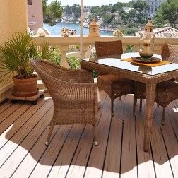 Wicker patio furniture on a wooden deck with ocean view. A glass-topped table and chairs, with potted plant.