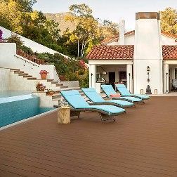 Poolside patio with blue lounge chairs on brown composite decking, villa in background.