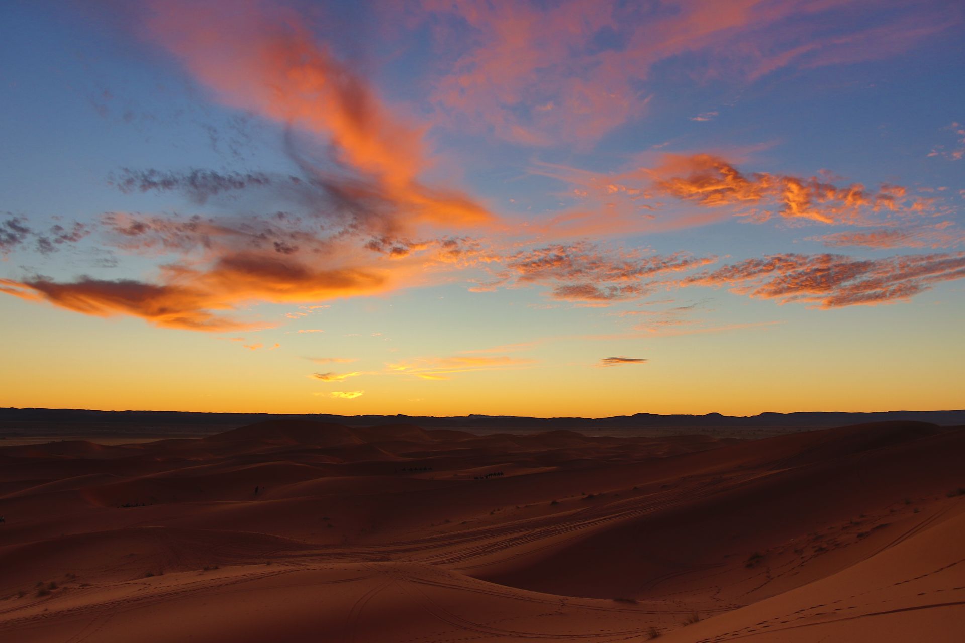 Sunset over sand dunes with orange-tinged clouds in a blue sky.