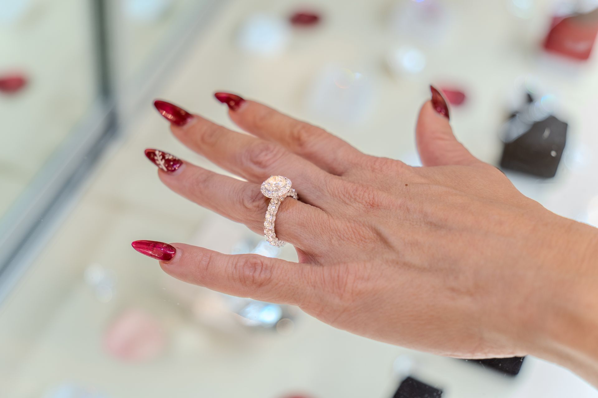 Hand with red nails, displaying a rose gold engagement ring with a halo diamond setting.