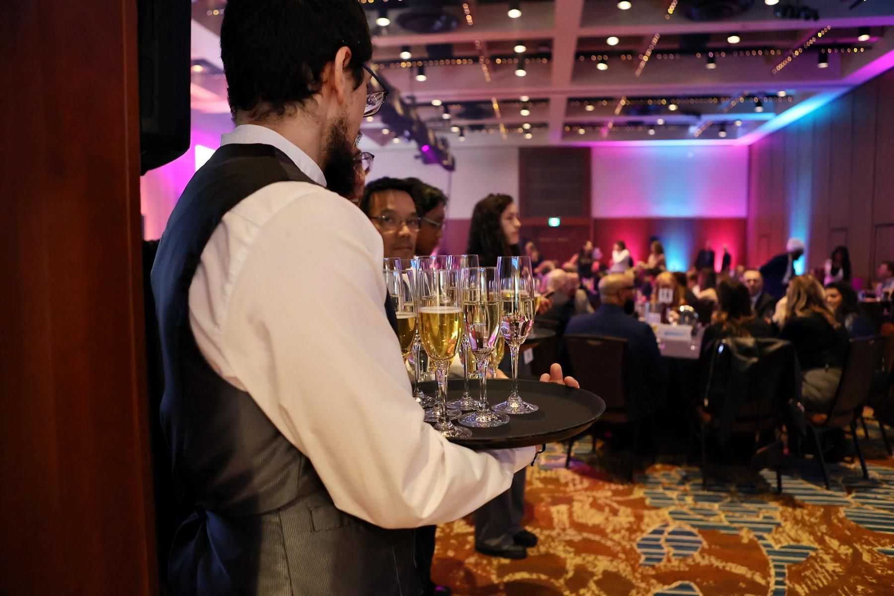 A waiter is carrying a tray of champagne glasses in a large room.