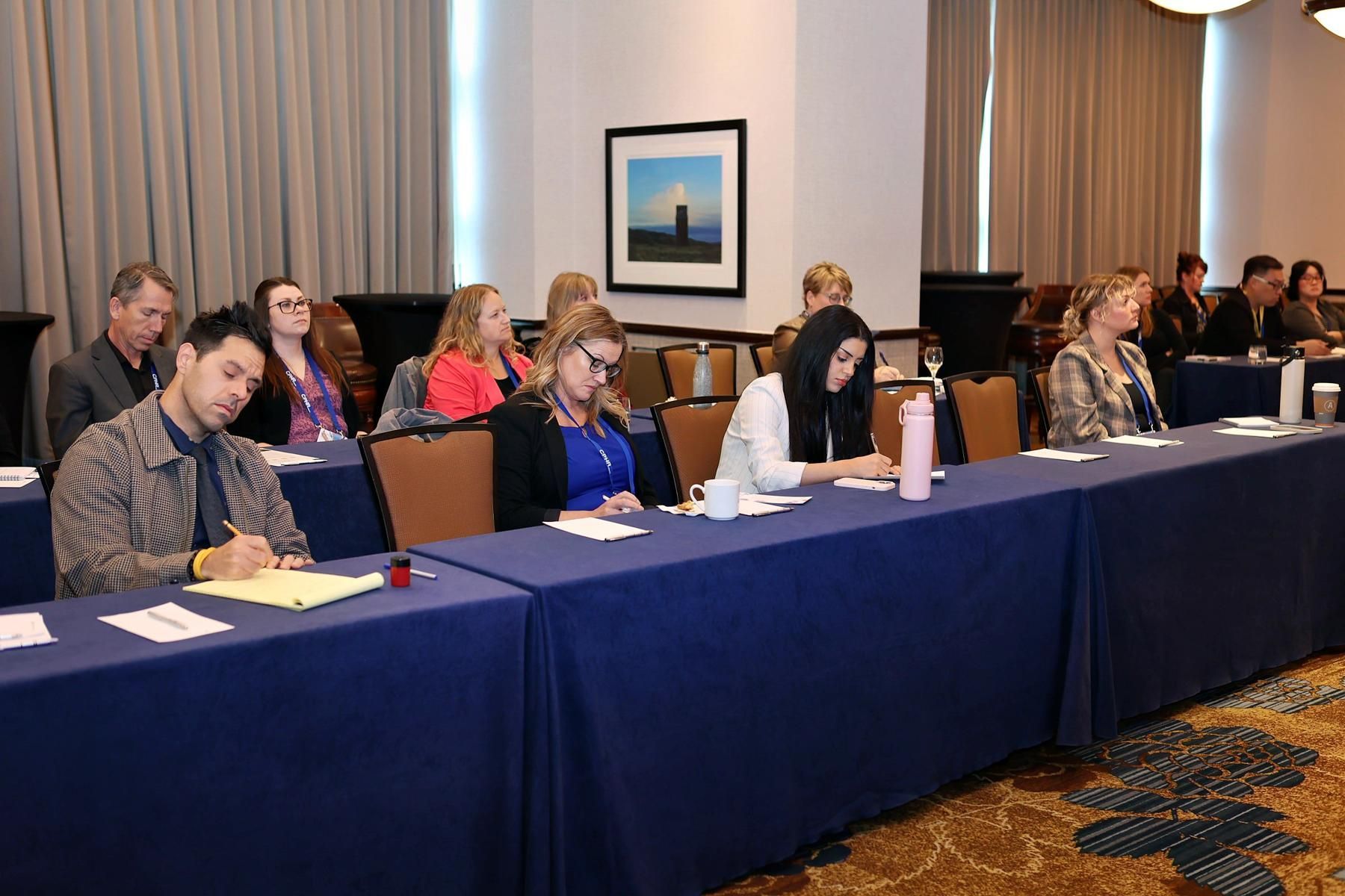 A group of people are sitting at long tables in a conference room.